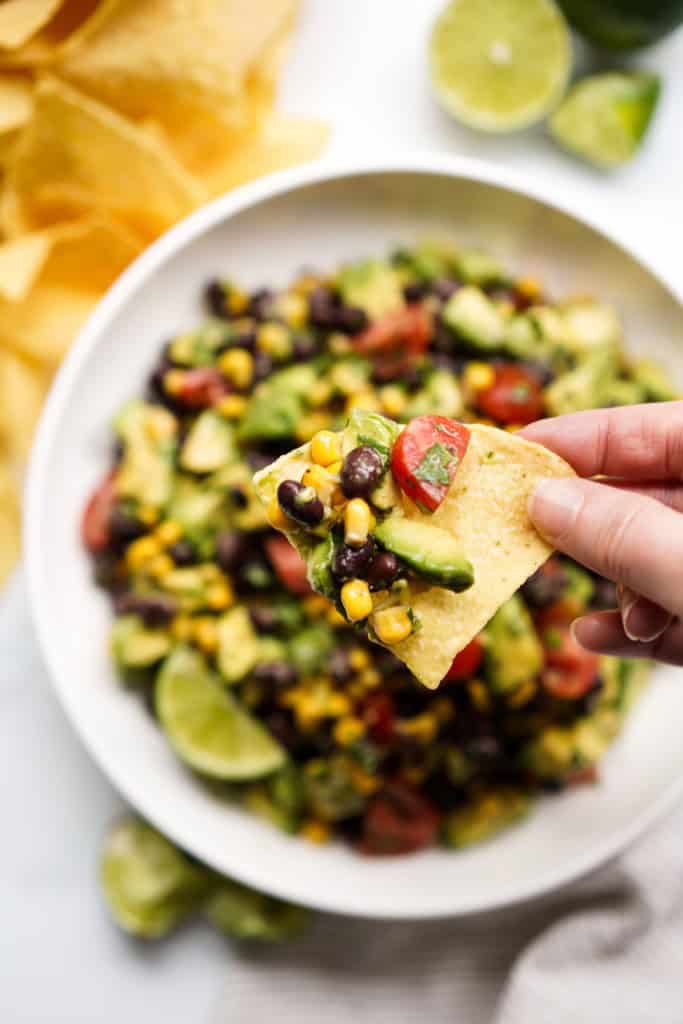 Lifting up a piece of tortilla chip topped with avocado corn salsa