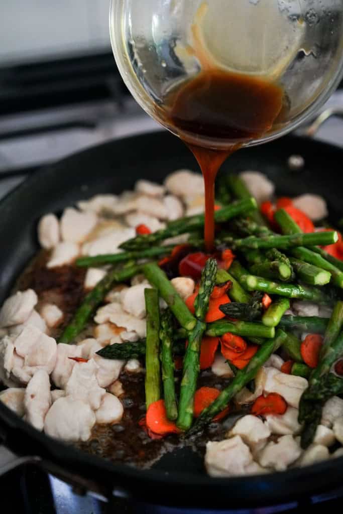pouring stir fry sauce into a skillet with chicken, asparagus and carrots