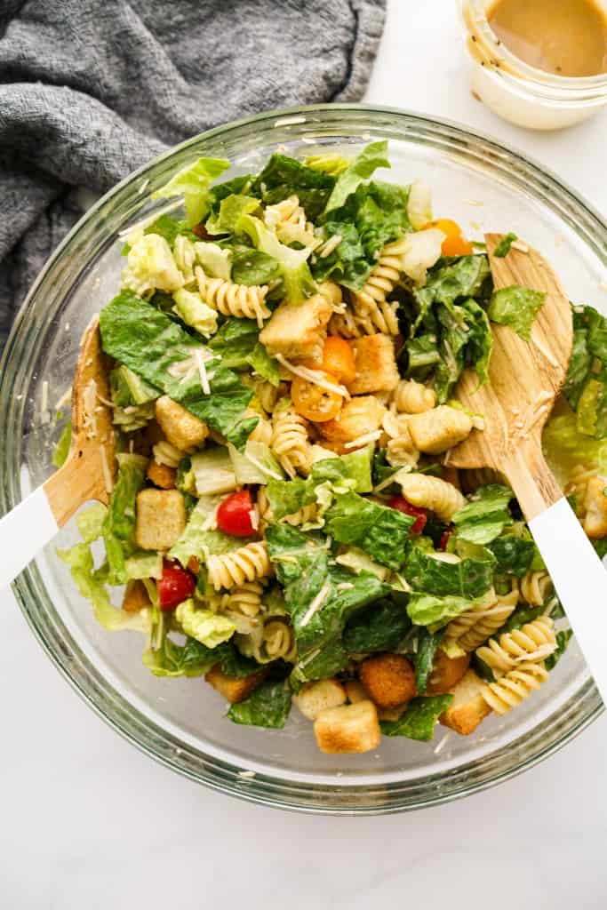 Tossing caesar pasta salad with dressing and croutons in a large mixing bowl, using salad spoons