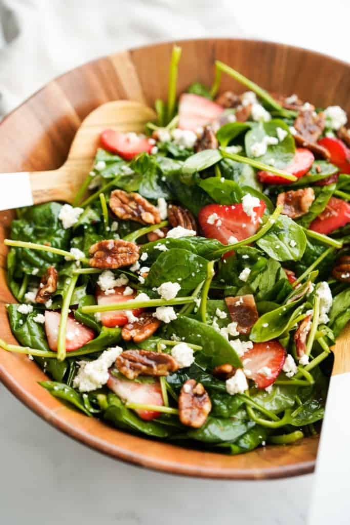 A large bowl of strawberry bacon spinach salad tossed in dressing, topped with pecans and cheese