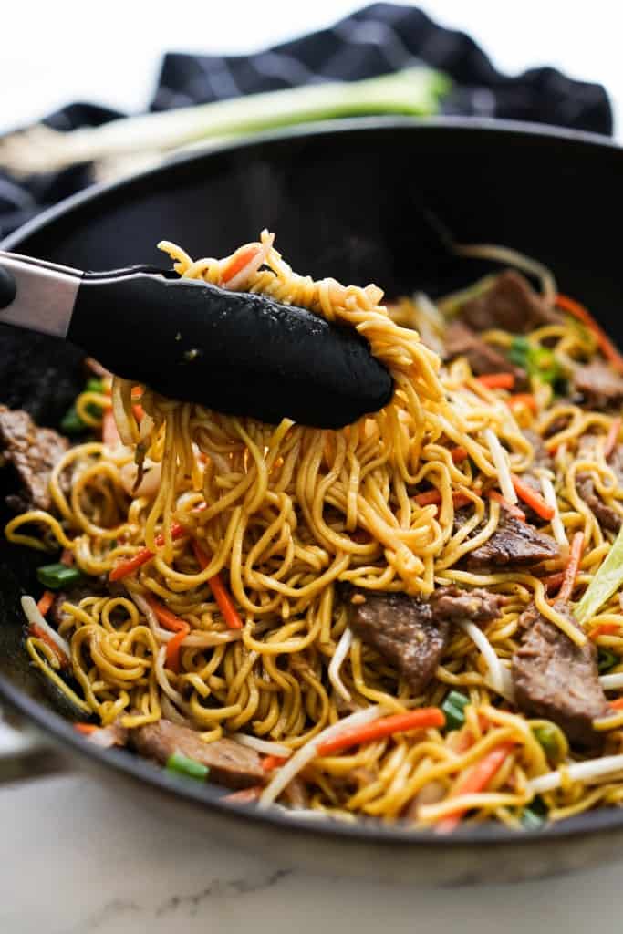 Lifting up a bunch of chow noodles from large wok filled with noodles, beef and veggies