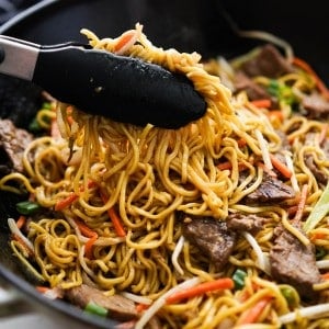 LIfting up beef chow mein noodles using a pair of tongs