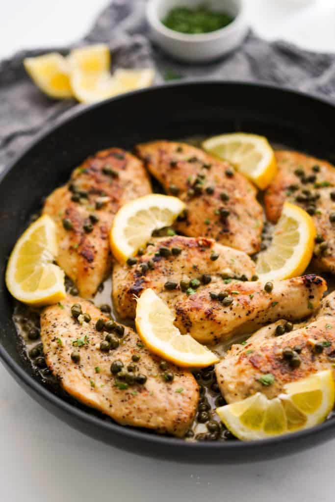 A skillet loaded with lemon chicken piccata in lemon caper sauce, with lemon wedges and a bowl of chopped parsley in the background