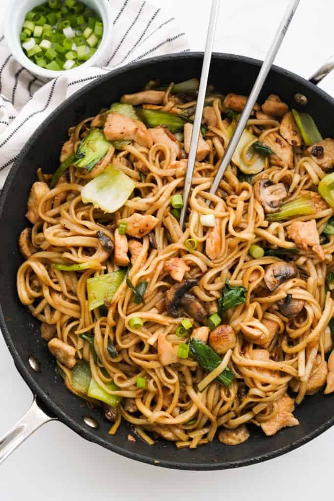 A large skillet loaded with udon noodles stir fried with bok choy, mushrooms and chicken