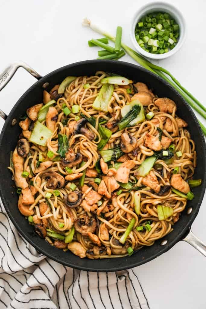 A large non stick skillet loaded with stir fried udon noodles, topped with chicken, mushrooms and bak choy