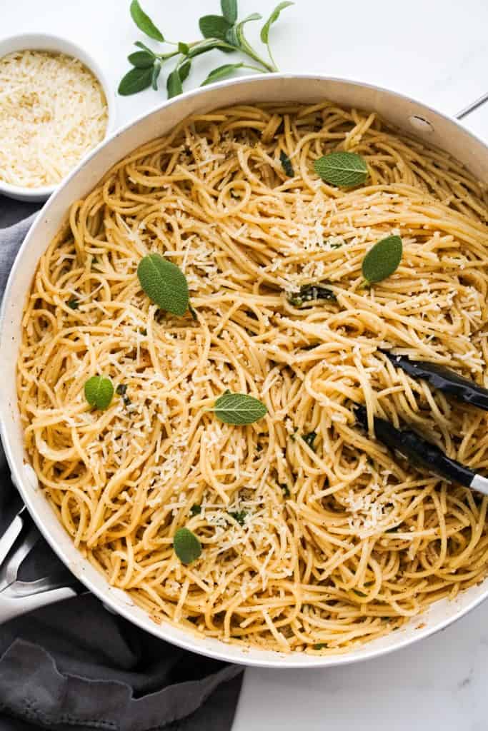 Brown butter pasta in a large skillet tossed in sage leaves, with a small bowl of parmesan cheese in the background