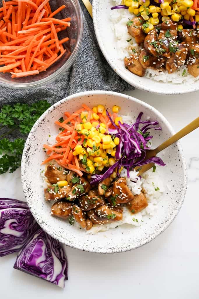 A bowl of rice topped with chicken with soy glaze, shredded carrots, corn and shredded cabbage