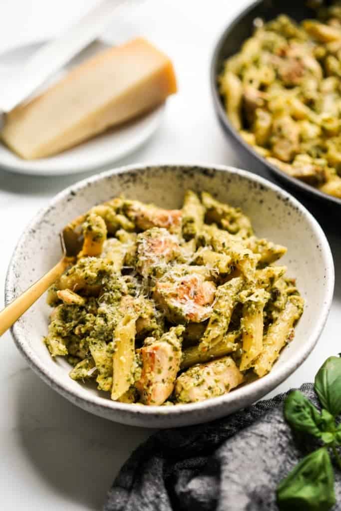 A serving bowl of creamy pesto chicken pasta, with a large skillet behind it