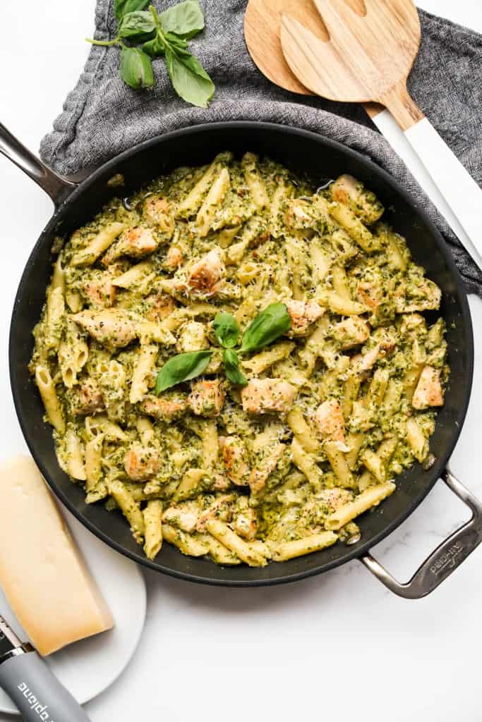 A large skillet loaded with creamy pesto pasta and chicken pieces, with fresh basil leaves and a block of parmesan cheese in the background