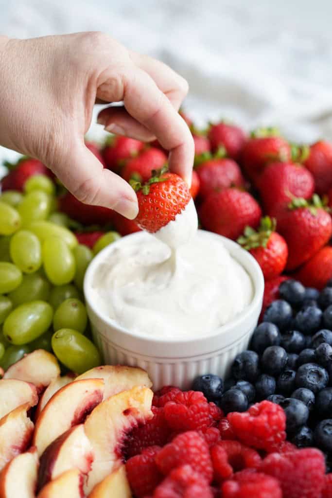 Dipping a strawberries into a bowl of fruit dip, with fruits surrounding it.