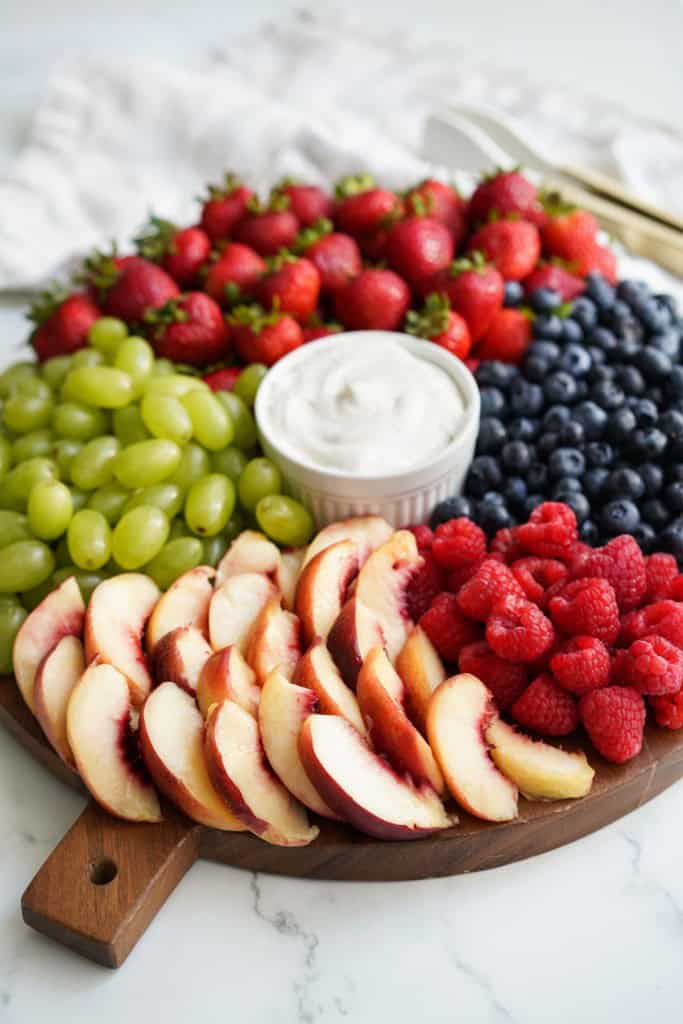 A large wooden round platter of fruits with a fruit dip in the center