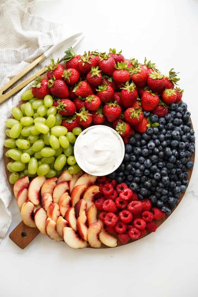 A round wooden board of strawberries, grapes, peaches, raspberries and blueberries with a bowl of marshmallow fruit dip in the middle