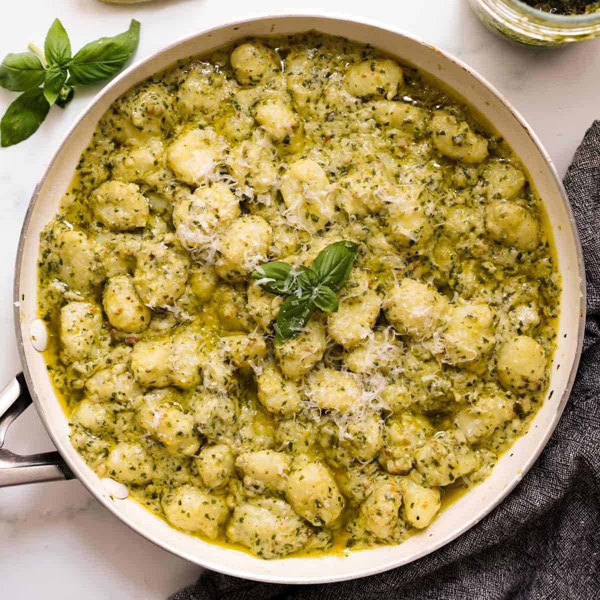 A skillet of gnocchi coated in creamy pesto sauce, topped with parmesan cheese and basil leaves